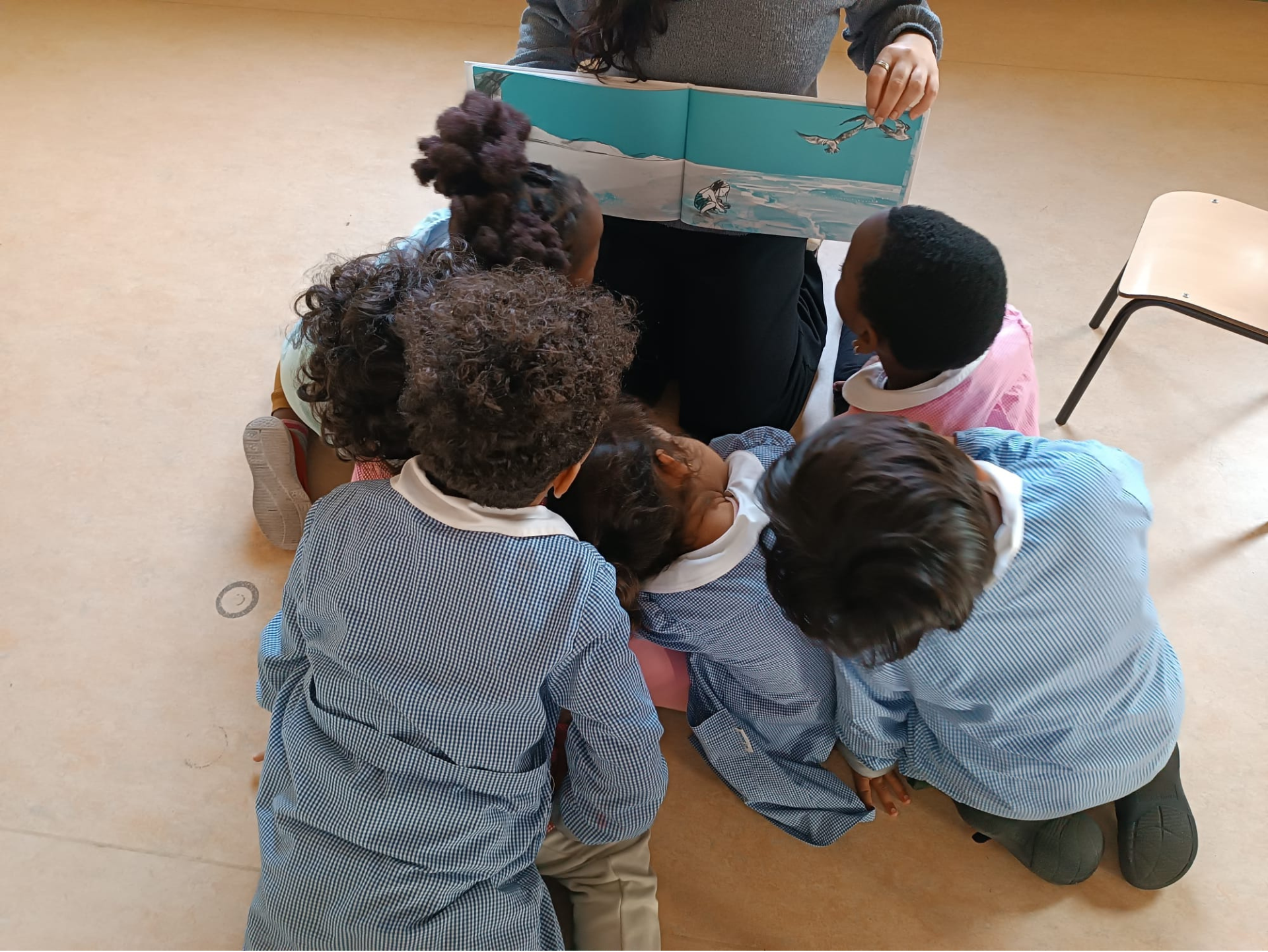 Copertina del laboratorio "Picturebook e silentbook" raffigurante un gruppo di bambini e bambine di spalle dal diverso background culturale a terra che guardano con interesse un libro illustrato "L'onda" che la maestra davanti a loro tiene aperto. La maestra è seduta sulle ginocchia.