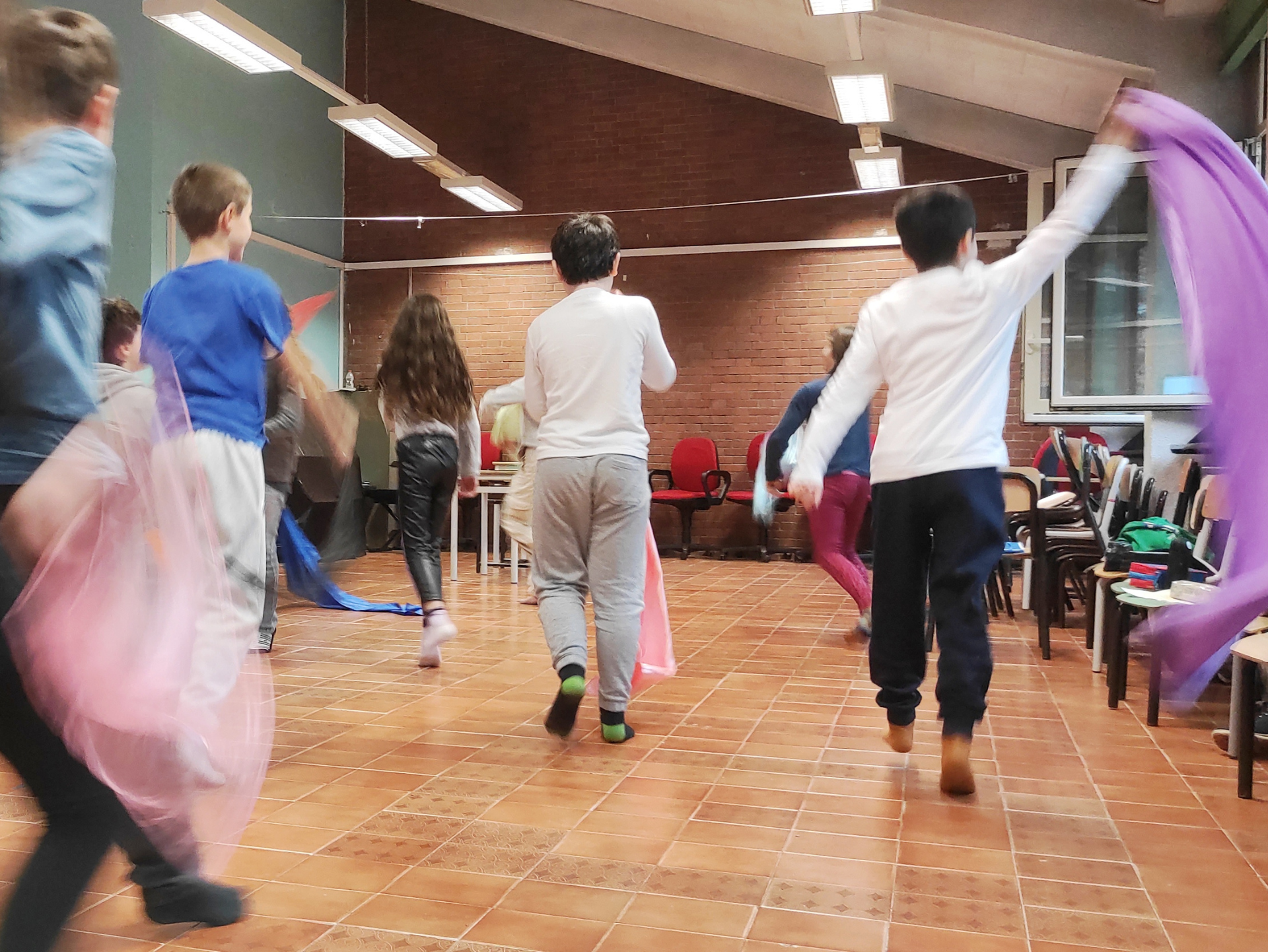 Copertina del laboratorio "Un mondo a colori - Educazione alla Interculturalità" raffigurante un gruppo di bambine e bambini tra i 6 e i 10 anni che si muovono per la classe con dei tessuti colorati. la foto è sfocata per mostrare la dinamicità e il movimento dell'attività